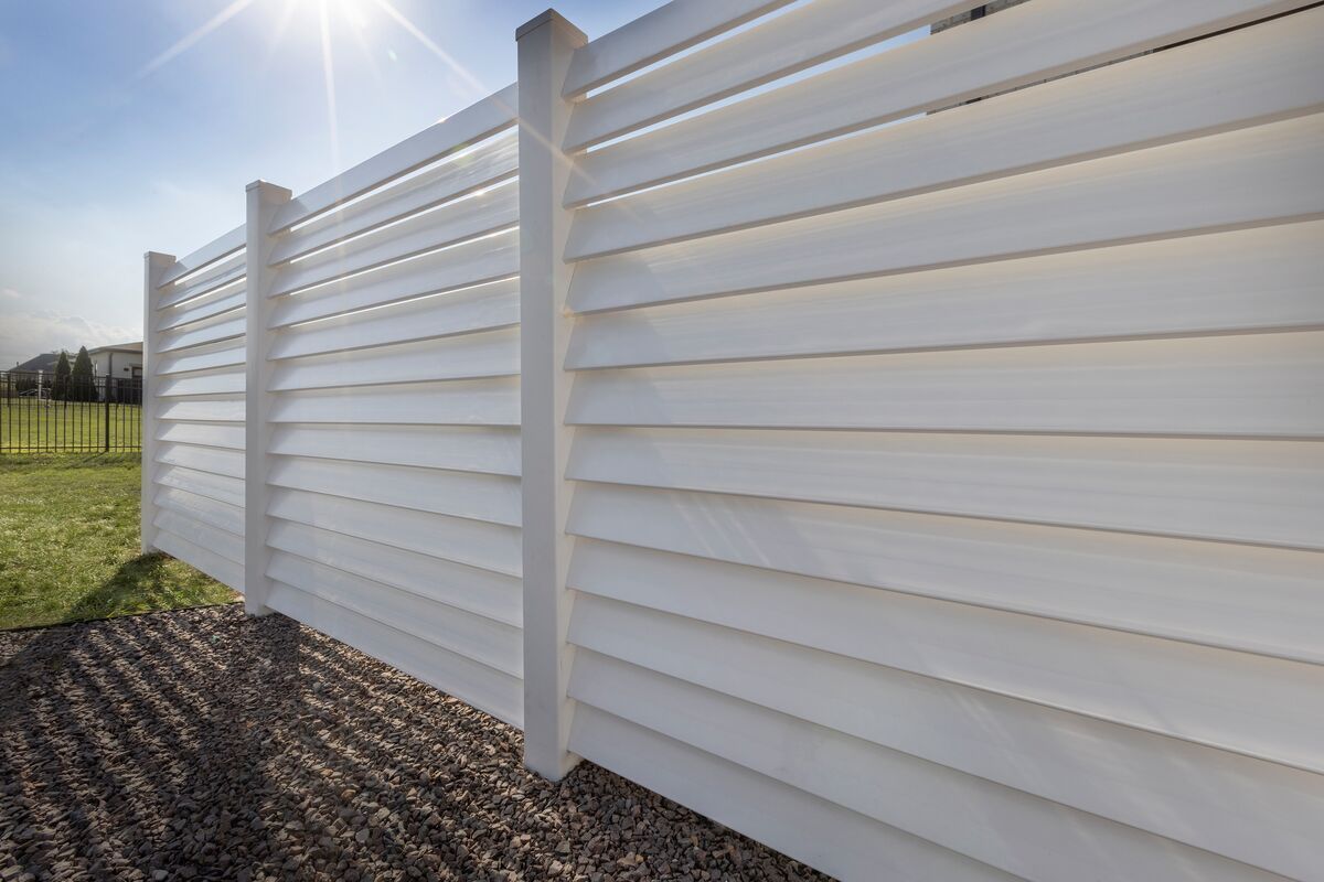 White slatted fence in sunlight, dividing a grassy yard from a gravel area.