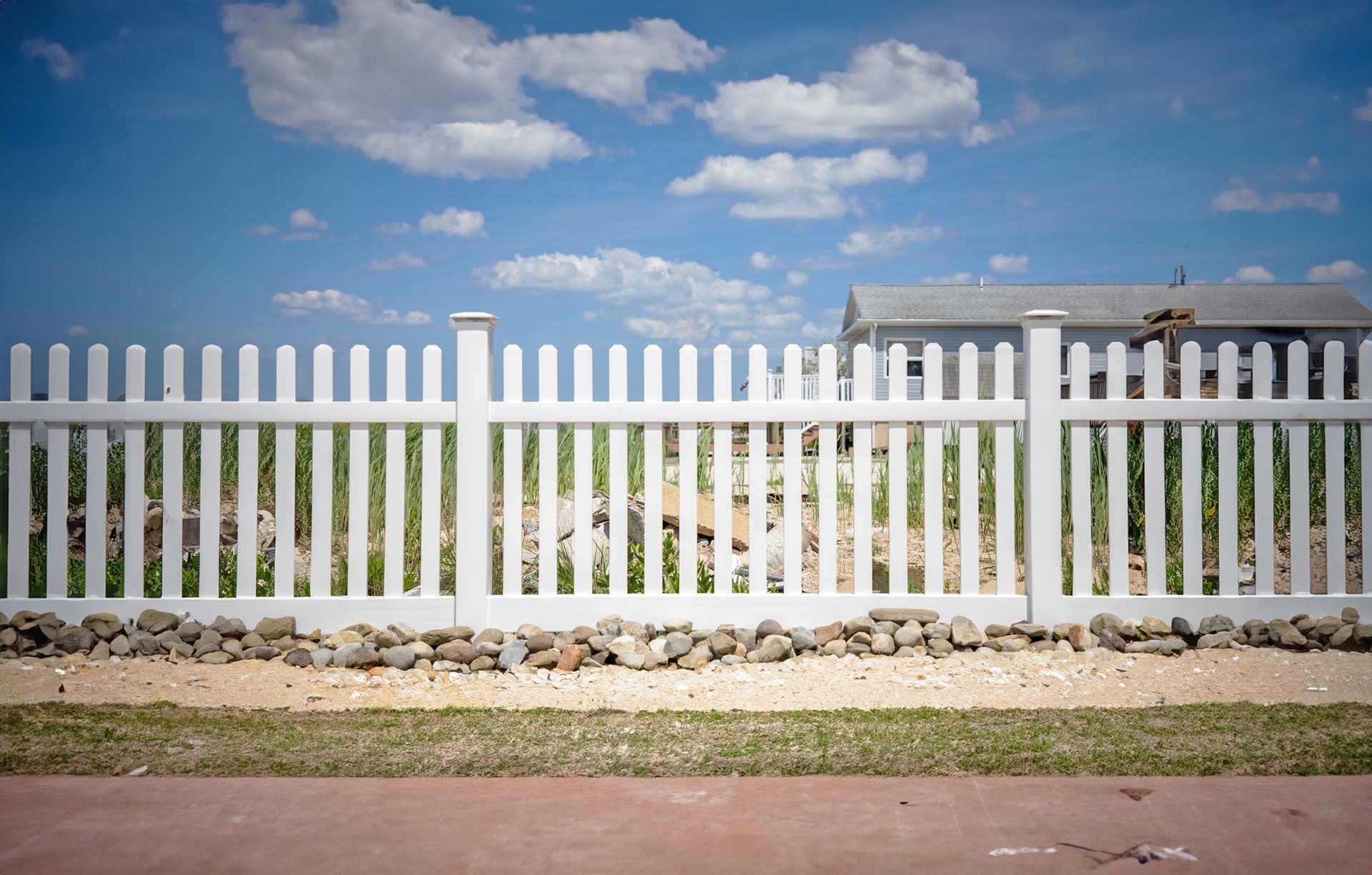 White picket fence in front of a house, set against a blue sky with fluffy clouds.