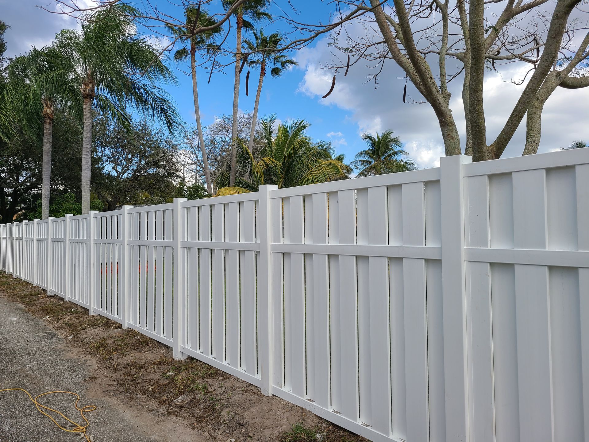 A white fence surrounds a dirt road with palm trees in the background.