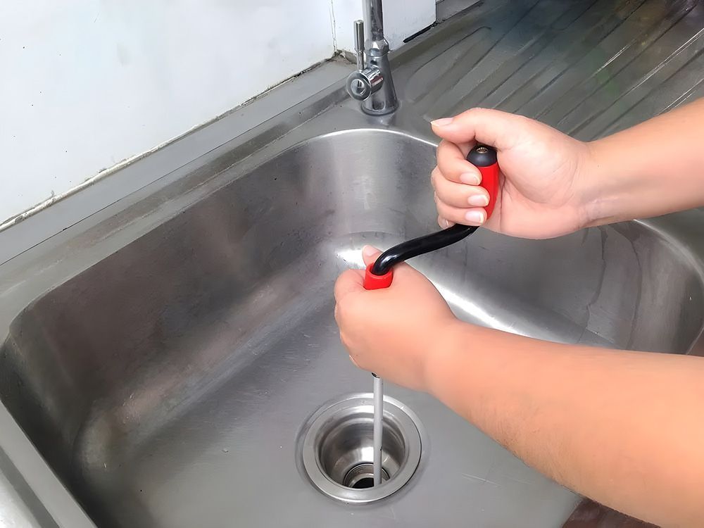 A person using a red-handled sink auger to clear a blockage in a stainless steel kitchen sink drain.