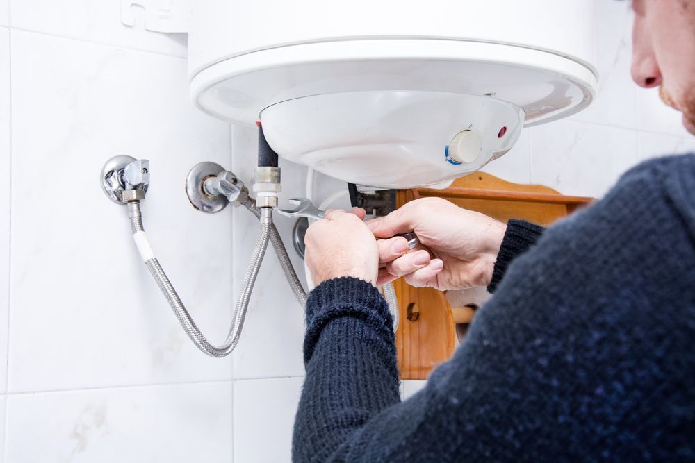 A person using a wrench to repair plumbing pipes connected to a white wall-mounted water heater.