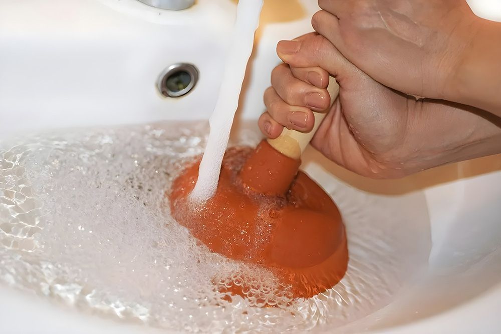 A person uses a red rubber plunger in a white bathroom sink filled with soapy water and a running faucet.