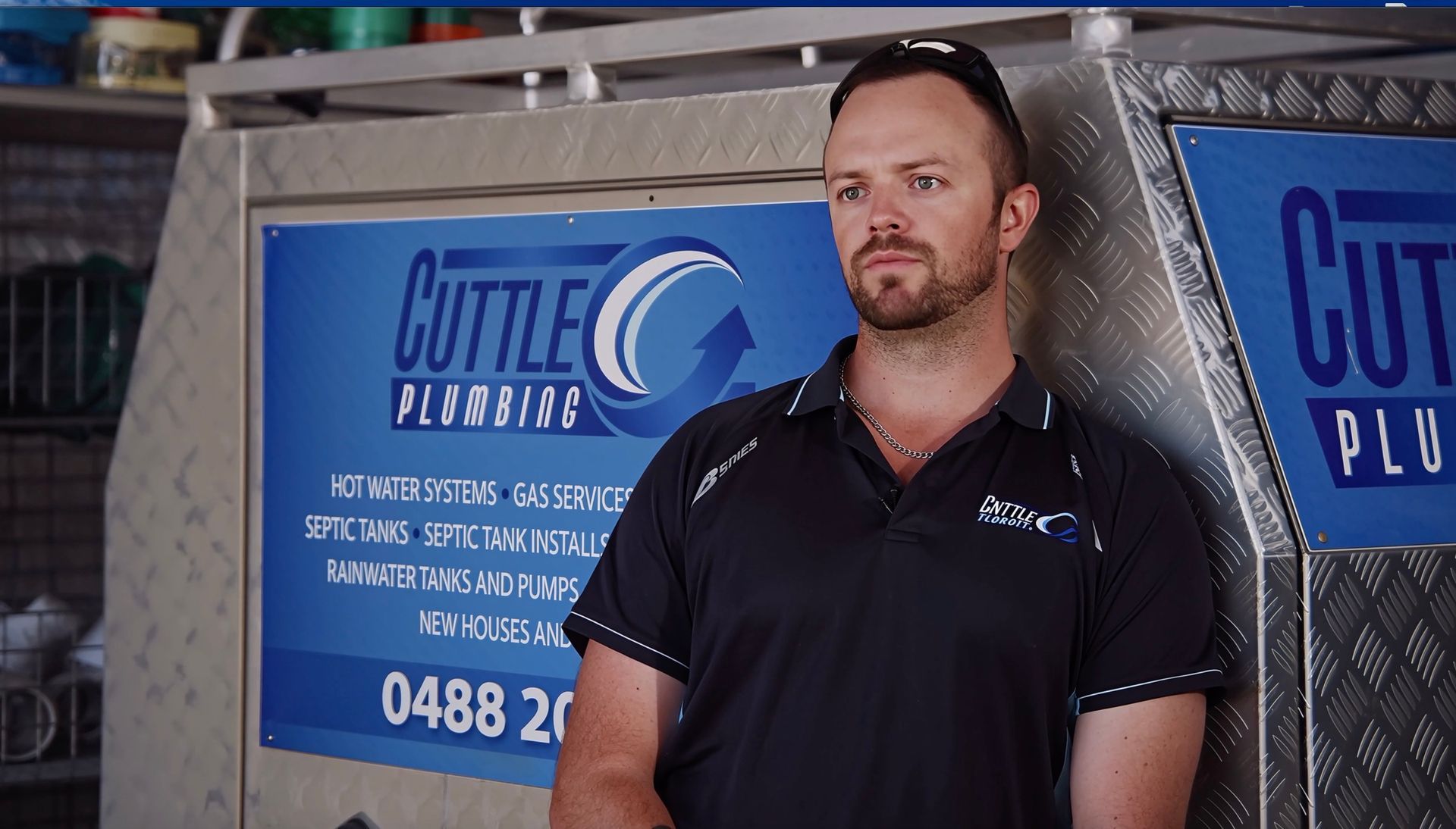 A man in a black work shirt leans against a Cuttle Plumbing service vehicle with a blue branded sign.