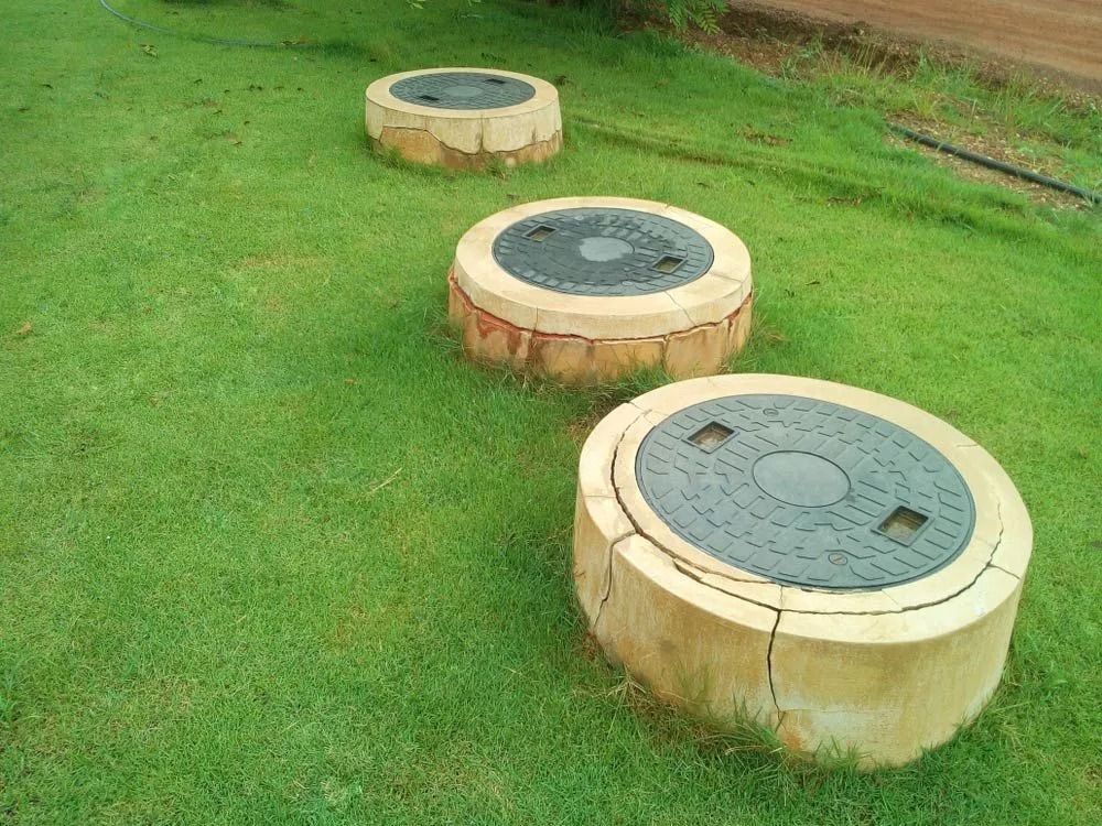 Three circular concrete maintenance hole covers with dark metal tops arranged in a line on a green grass lawn.