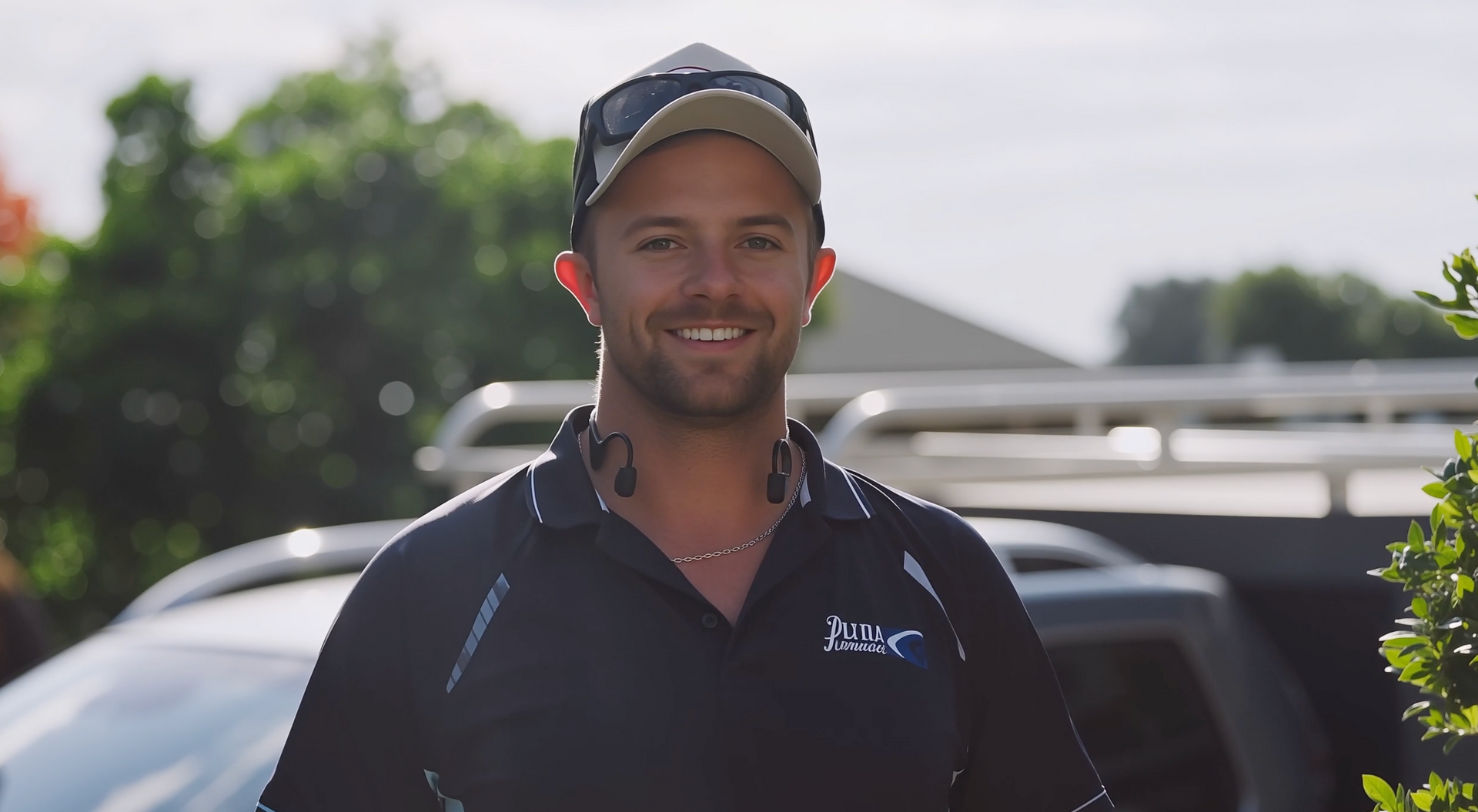 A person smiling outdoors wearing a baseball cap and dark polo shirt, with a vehicle visible in the blurred background.