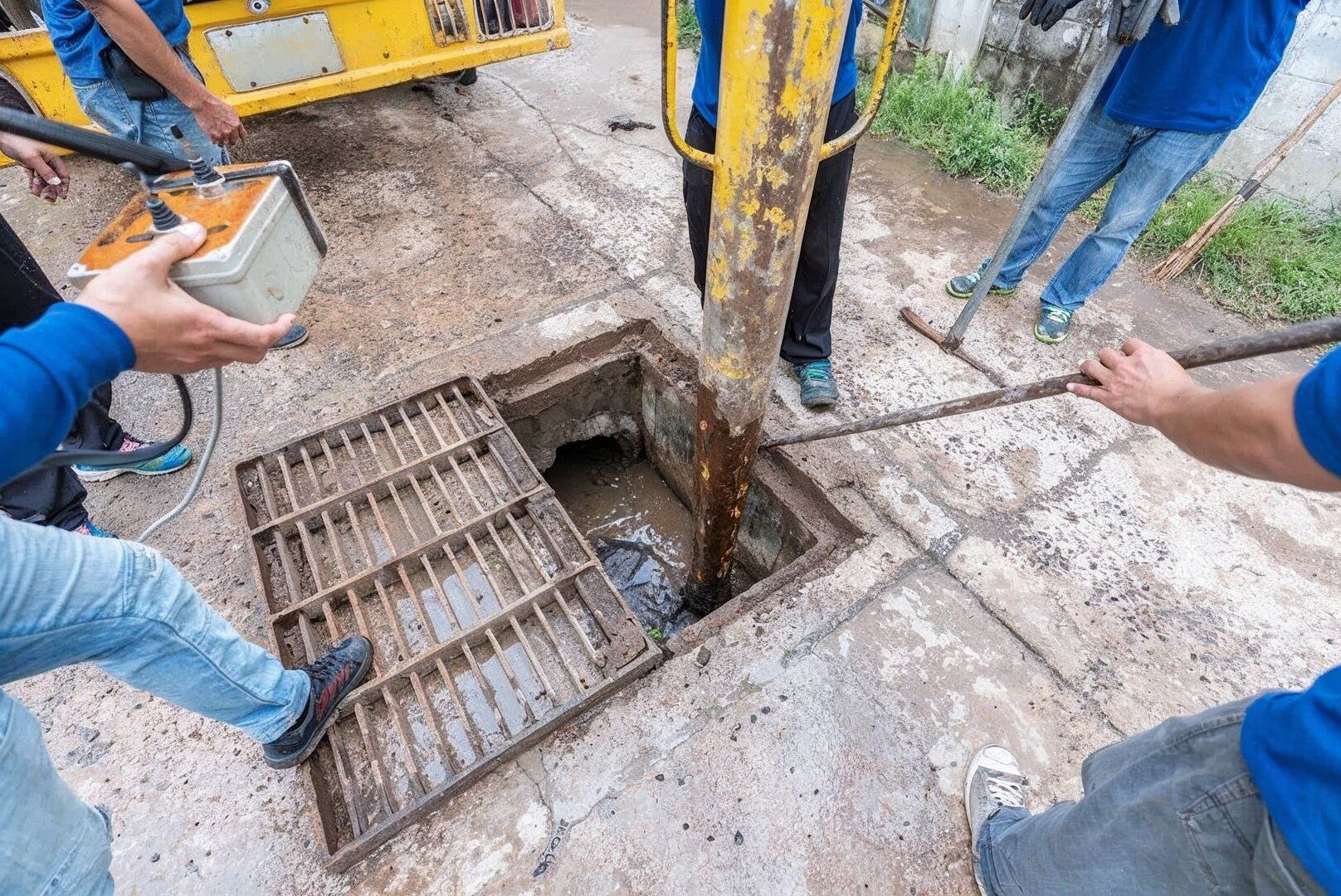 A group of people wearing blue shirts use tools to inspect or clean an open roadside storm drain.