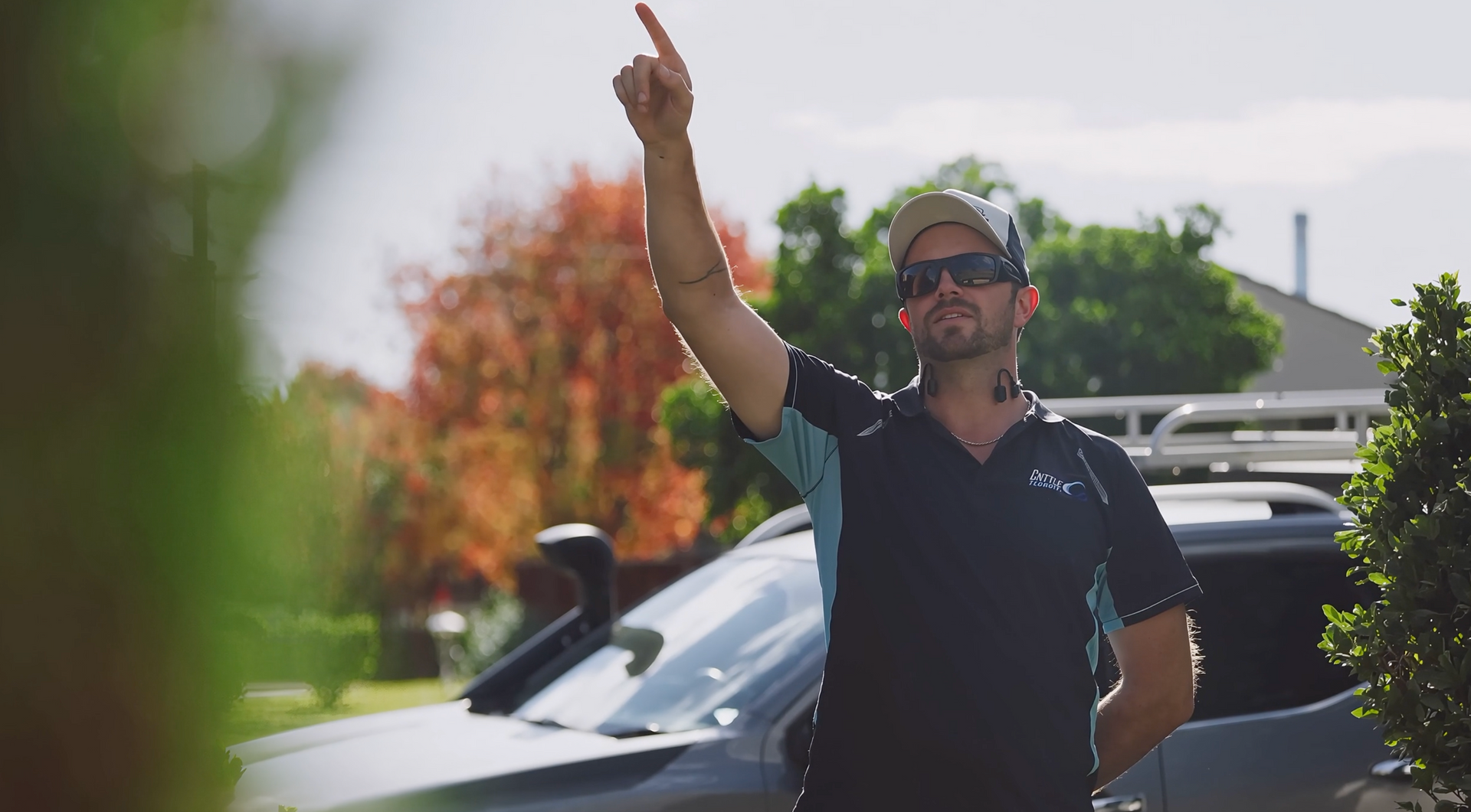 A person wearing sunglasses and a cap points upward while standing outdoors near a vehicle.