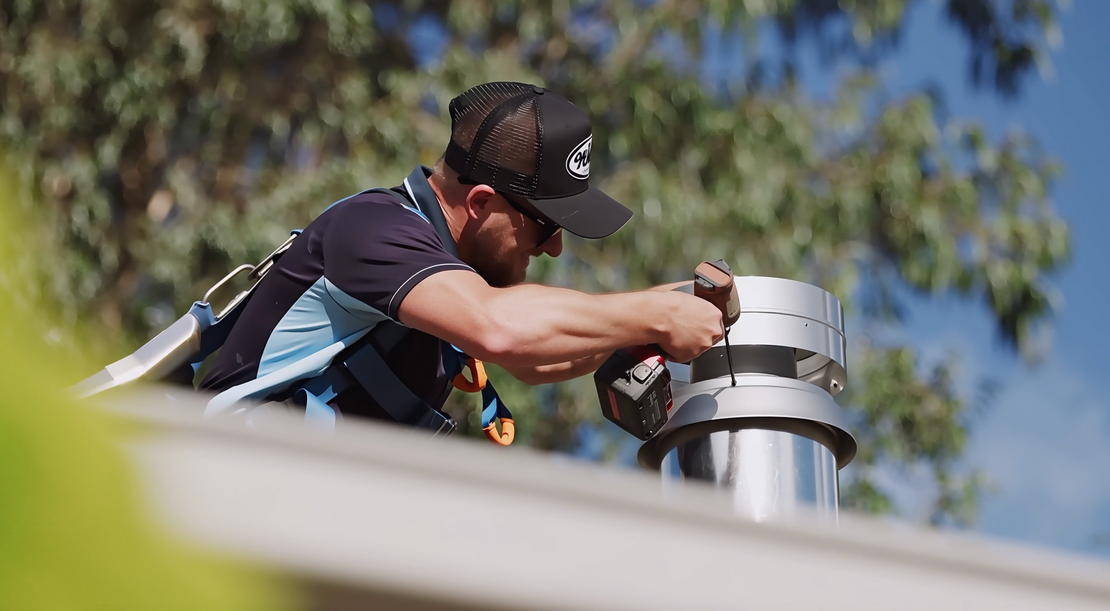 A worker wearing a safety harness and cap uses a power drill on a metal chimney flue on a rooftop outdoors.