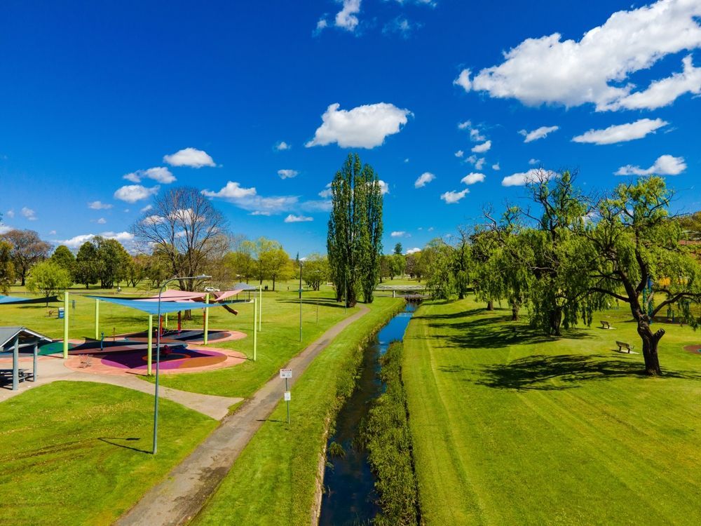 A park with a playground, a winding stream, a dirt path, and green trees under a bright blue sky with white clouds.
