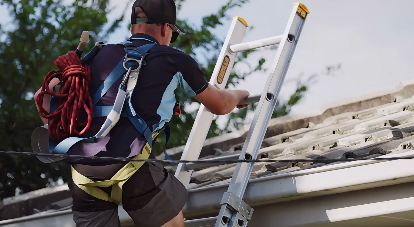 A person wearing a safety harness and carrying a coiled red rope climbs a ladder against a tiled roof.