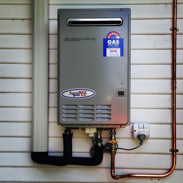 A gray AquaMAX gas water heater unit mounted on a white exterior wall with copper pipes and a plugged-in power cord.
