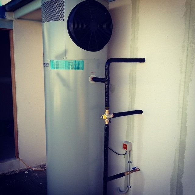 A silver cylindrical heat pump water heater sits against a white wall with black insulated pipes and electrical wiring.