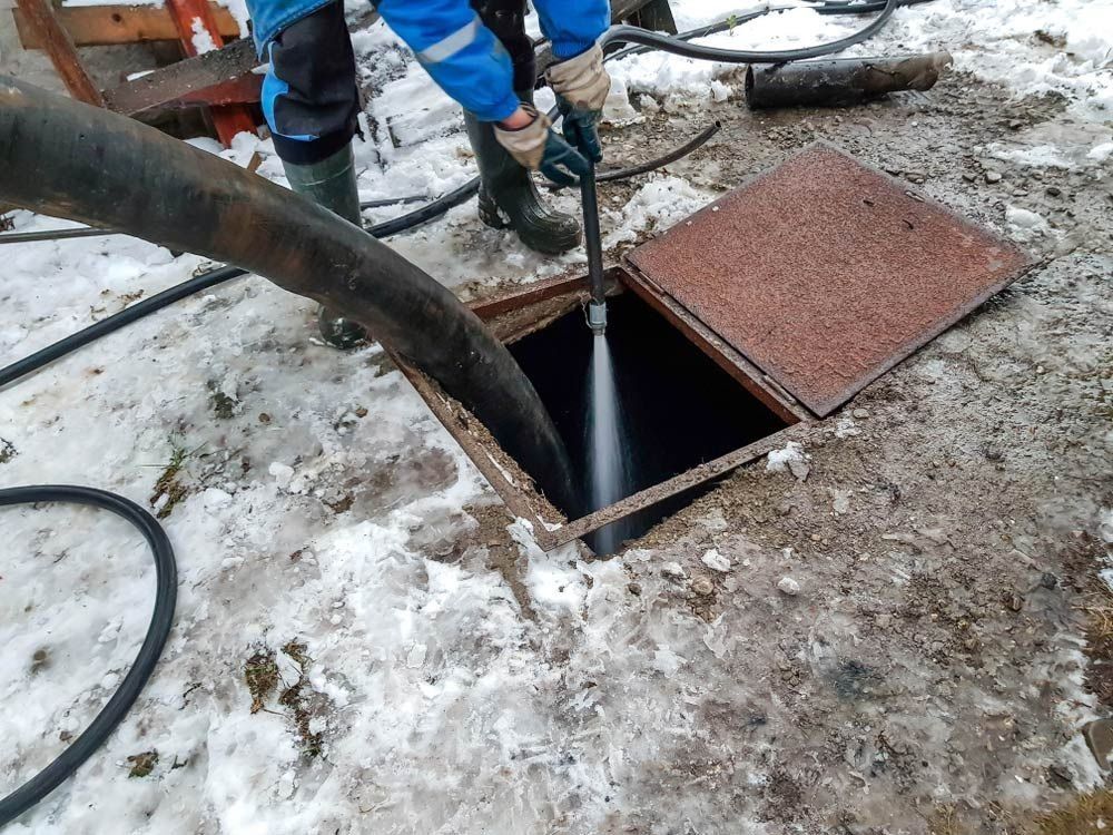 A worker in a blue uniform uses a high-pressure nozzle to spray water into an open underground maintenance hatch.