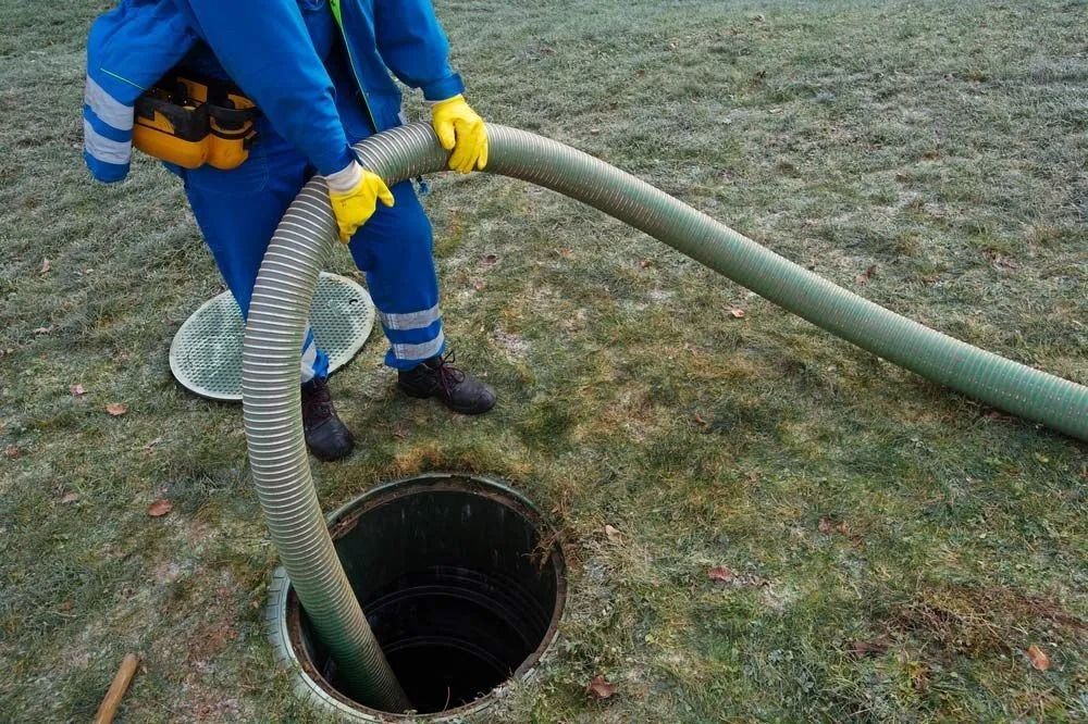 A worker in blue coveralls and yellow gloves feeds a large, flexible hose into an open manhole on a grassy lawn.