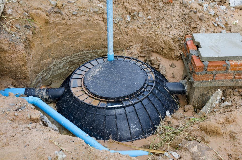 A black plastic septic tank is partially buried in an excavated dirt trench, connected to blue PVC drainage pipes.