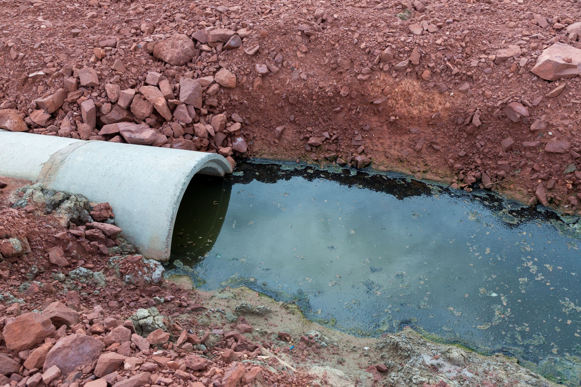 A concrete drainage pipe empties murky, dark water into a muddy trench surrounded by loose reddish-brown soil.