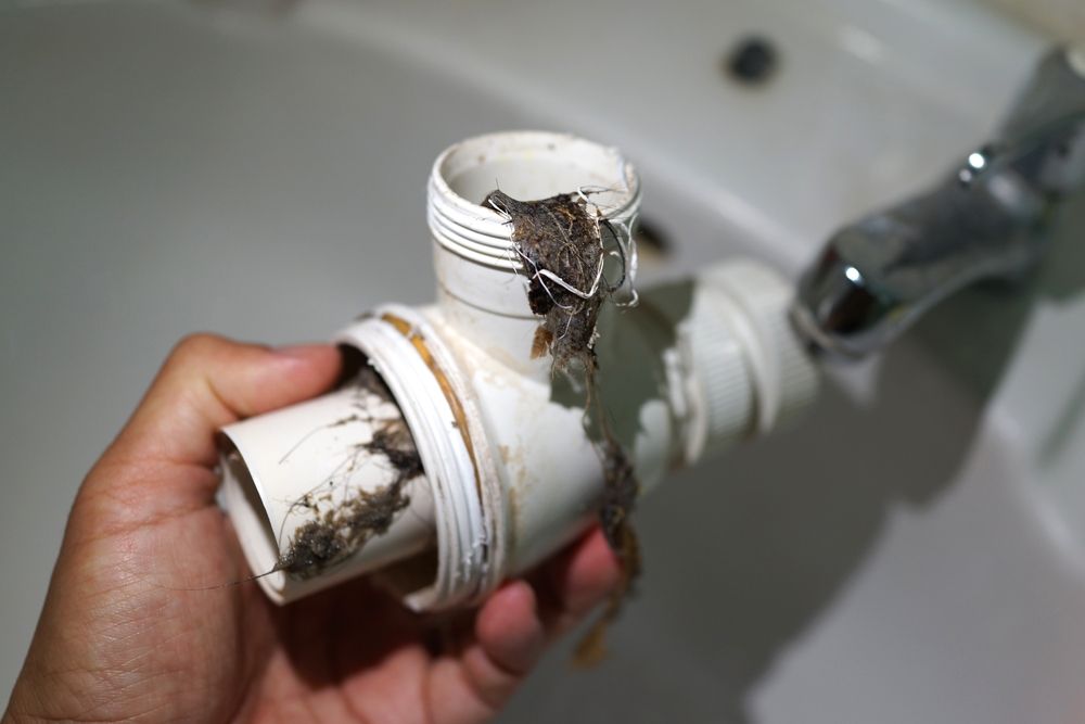 A hand holding a detached, clogged plastic sink pipe trap filled with hair and debris in a bathroom.
