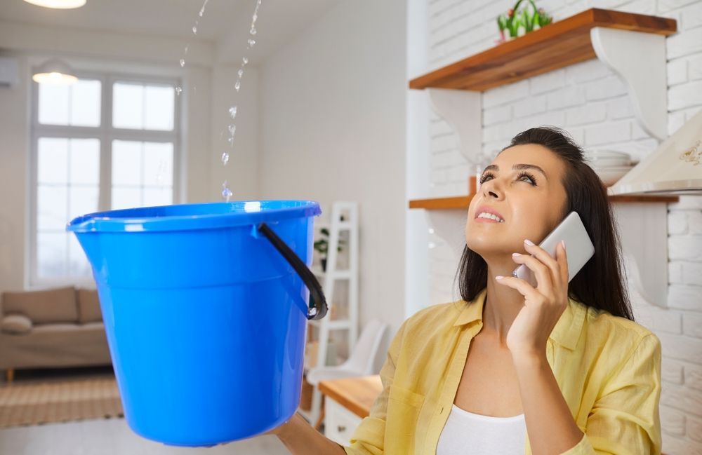 A person in a yellow shirt holds a blue bucket under a ceiling leak while speaking on a smartphone in a kitchen.