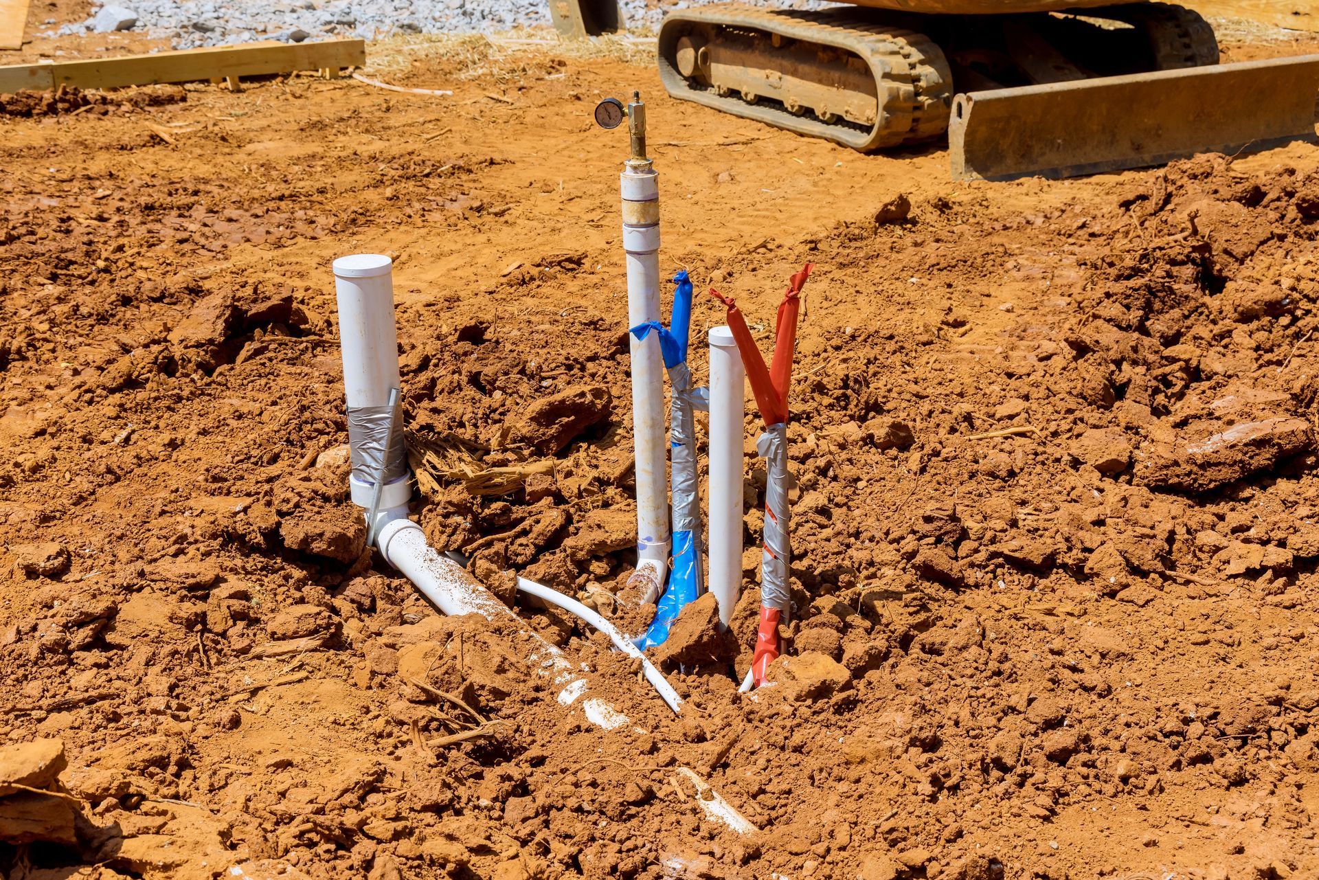 Plumbing pipes and conduits protruding from red dirt at a construction site, with a tractor track in the background.