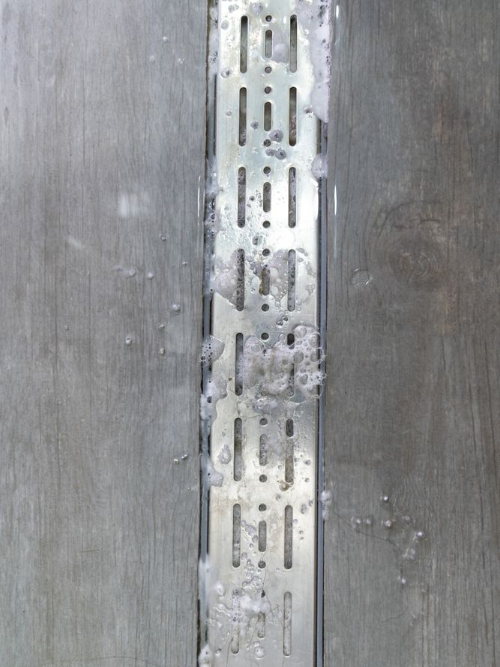 A metal linear shower drain embedded between grey, wood-look floor tiles with soapy water and bubbles on the surface.