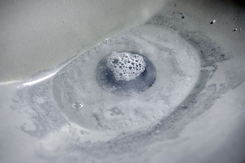 A white bathroom sink drain filled with grey, soapy water and a small patch of white bubbles.