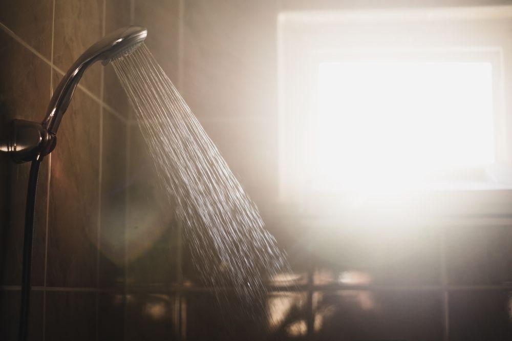 A shower head sprays water in a stream, illuminated by bright sunlight coming through a nearby window.