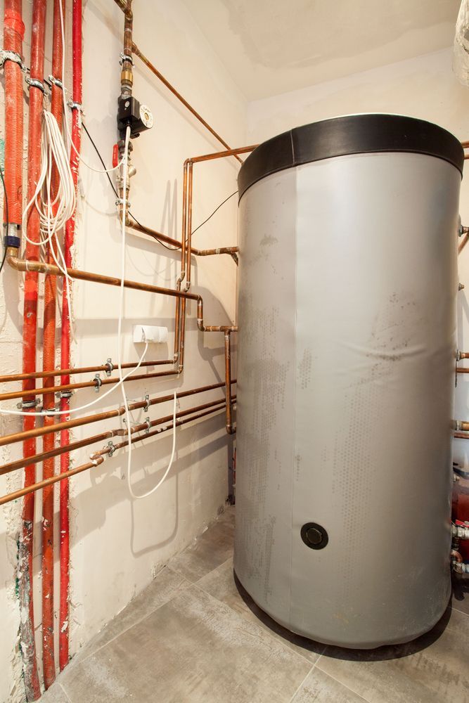 Large gray water heater tank in a utility room with copper pipes and red vertical conduits on the left wall.
