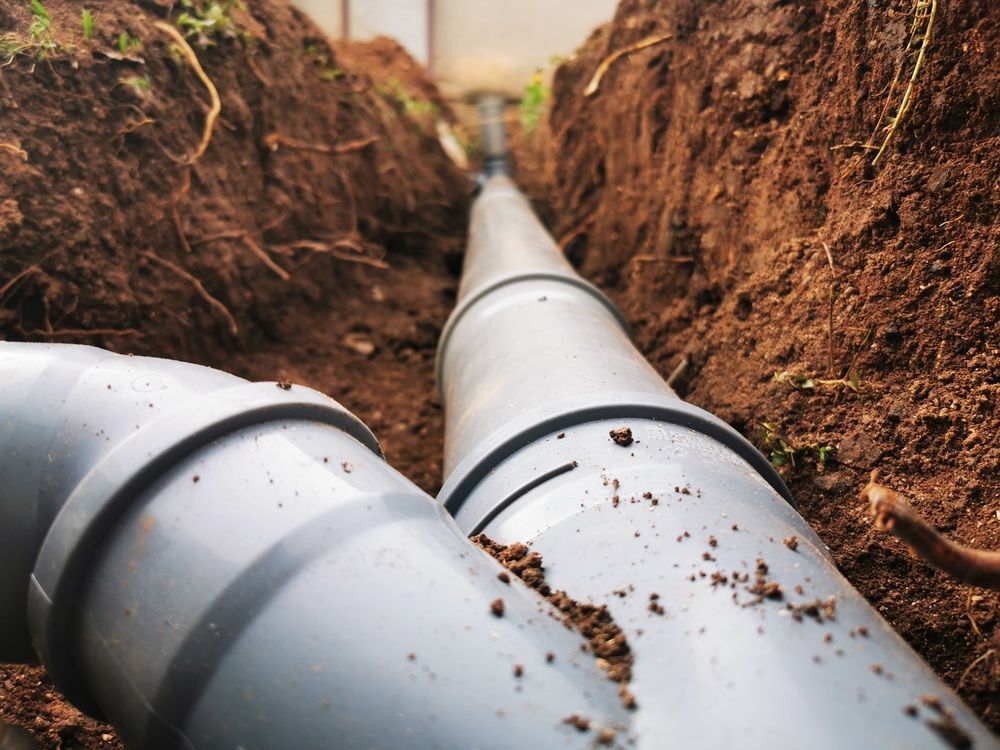 A grey plastic drainage pipe connection resting in a narrow, freshly dug dirt trench.