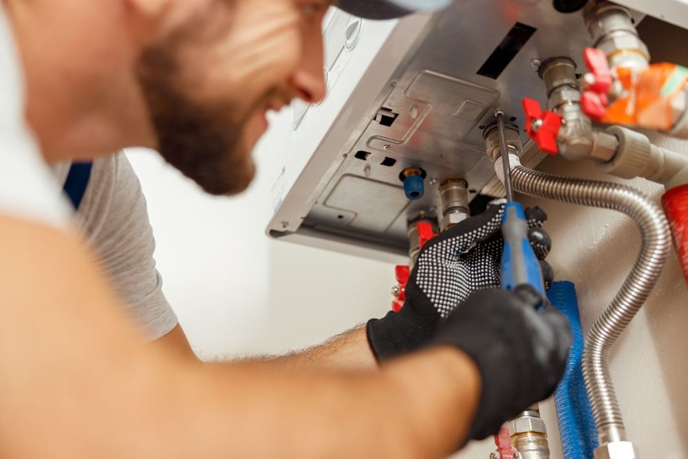 A technician in black gloves uses a screwdriver to repair a wall-mounted boiler with exposed pipes.