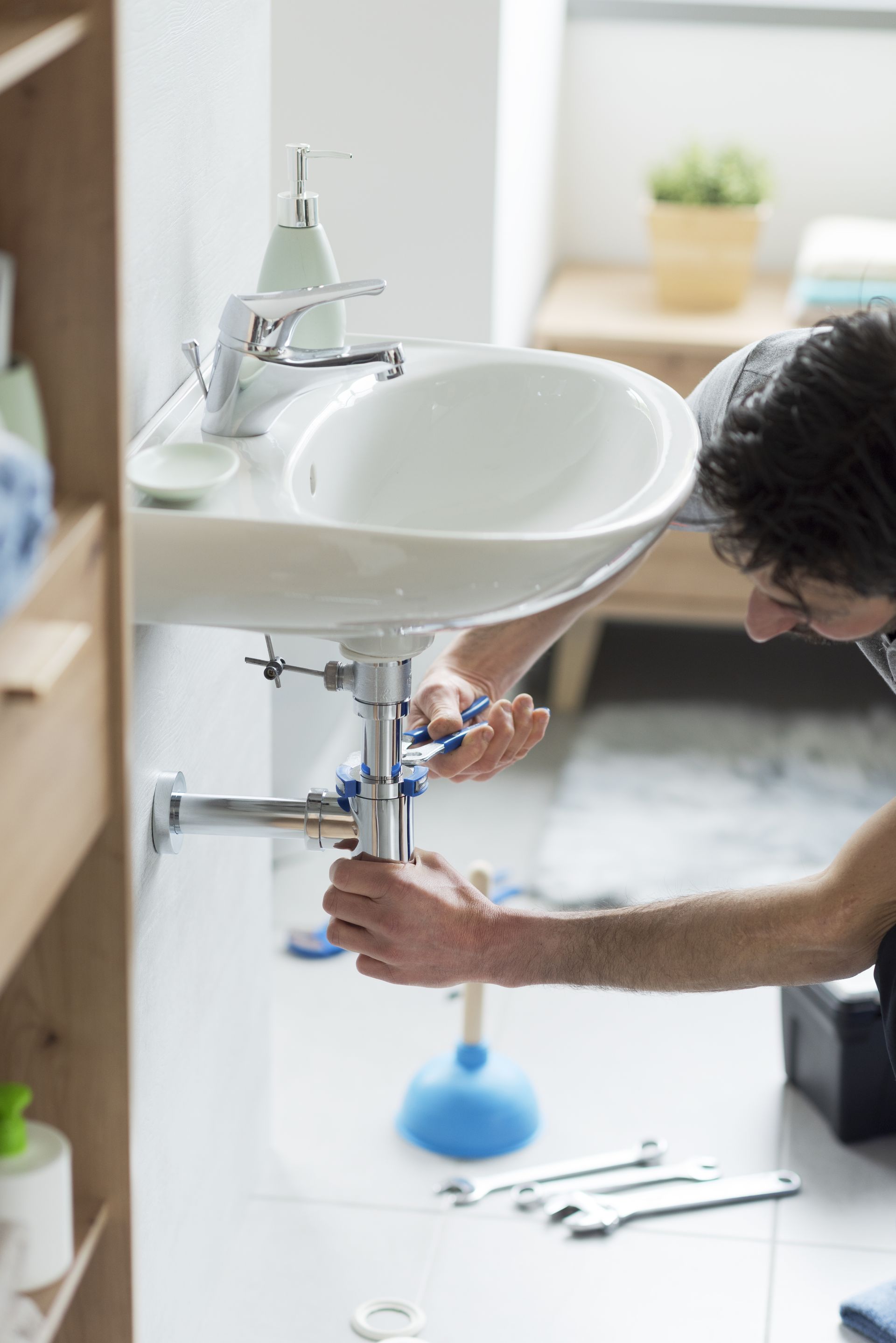 A person using a wrench to repair the metal pipes underneath a bathroom sink, with tools placed on the floor nearby.