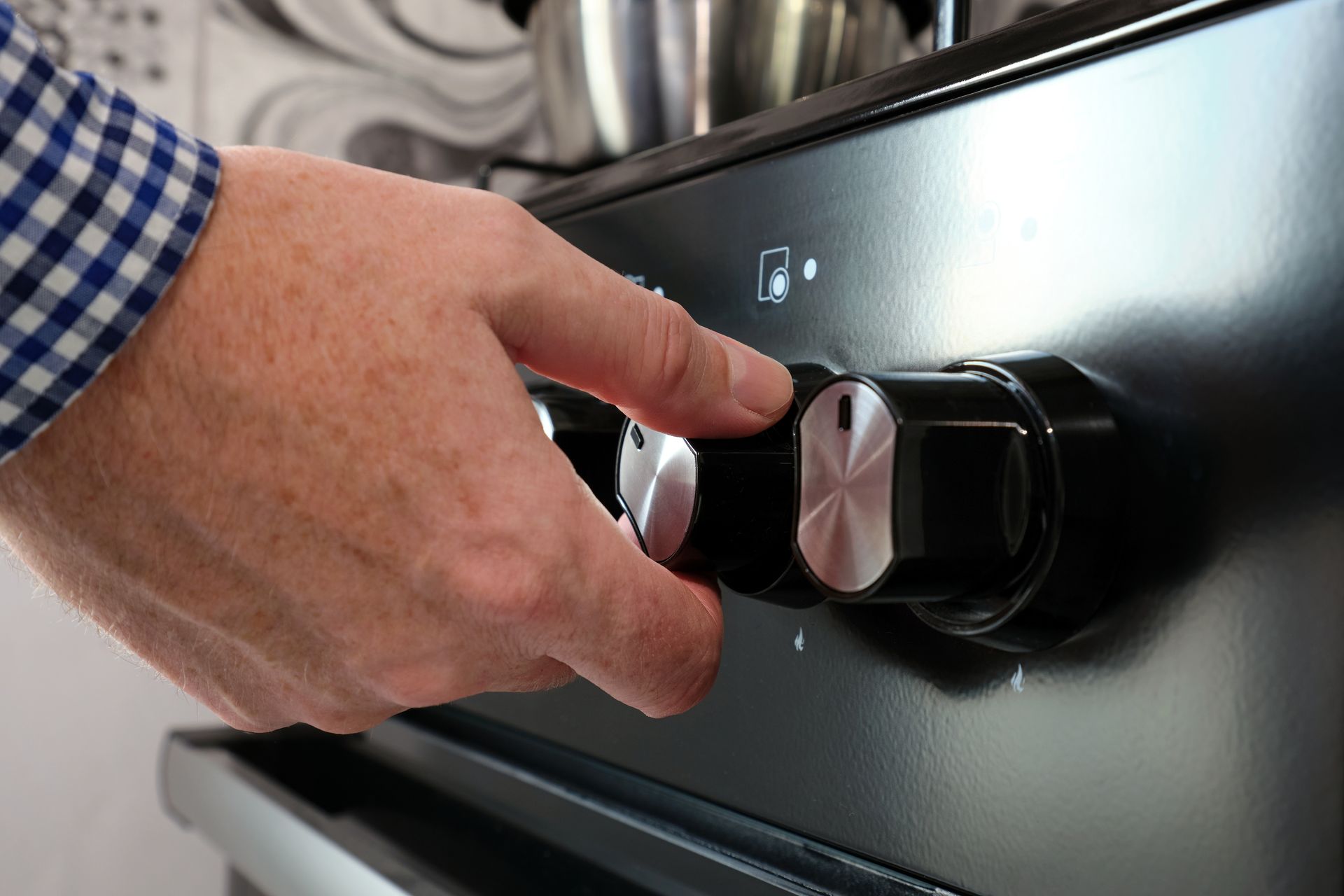 A hand in a blue checkered sleeve turns a black-and-silver control knob on a stove.
