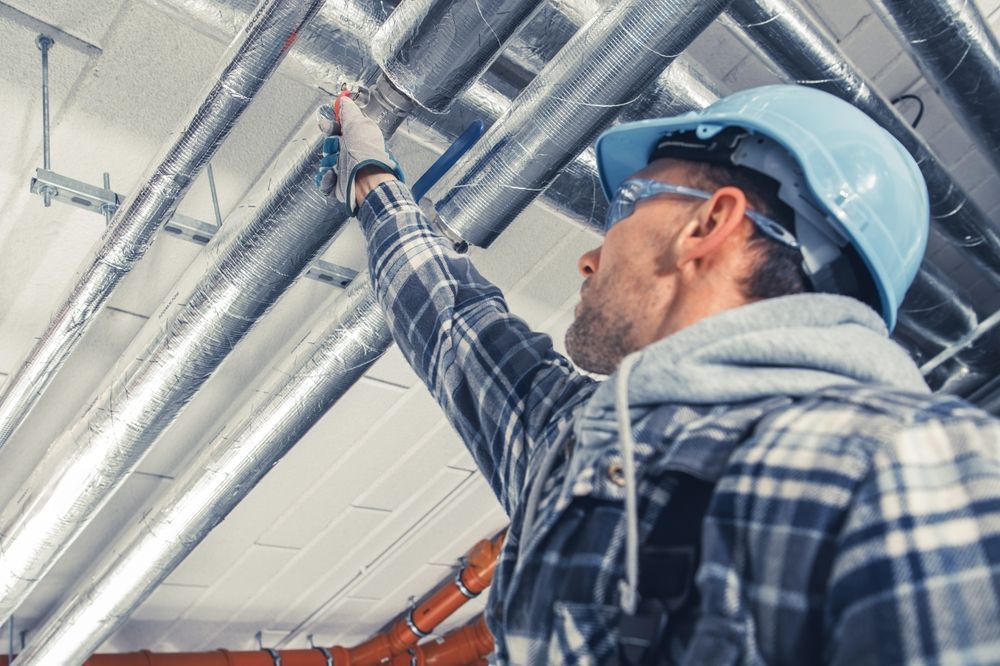 A construction worker in a blue hard hat and plaid shirt reaches up to inspect metallic pipes on a ceiling.