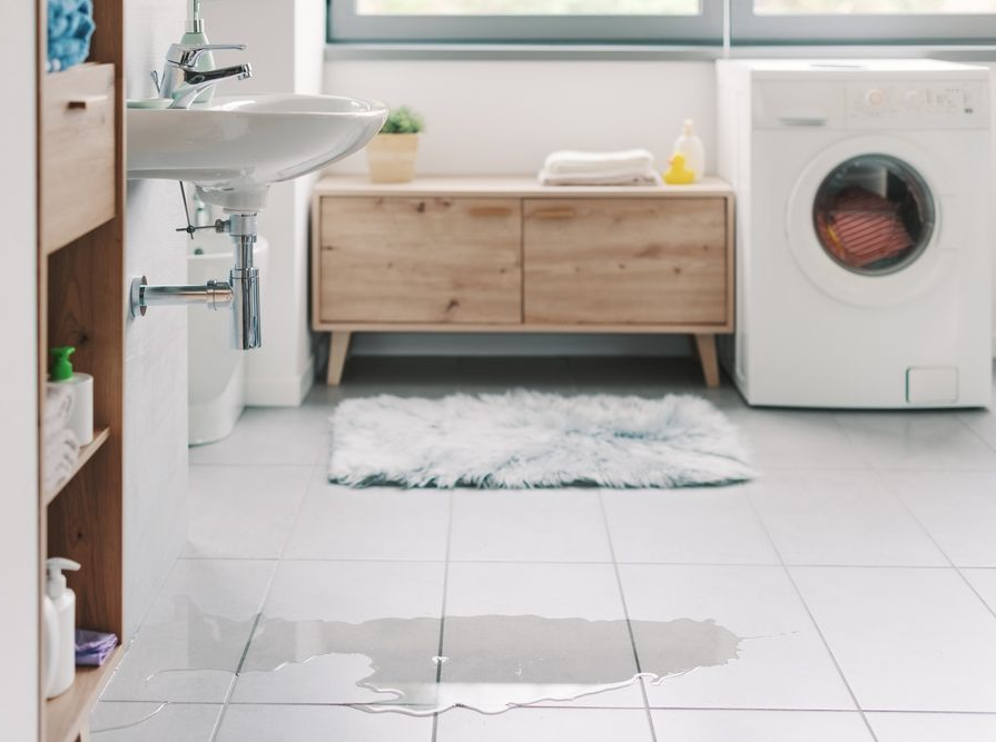 A water leak on the tile floor of a bright, modern bathroom featuring a sink and a front-loading washing machine.