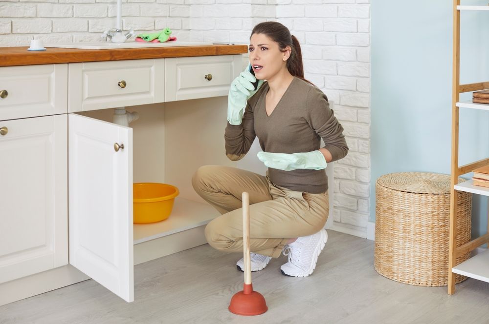 A person kneeling in a kitchen, wearing gloves and holding a phone while next to a plunger and an open cabinet.