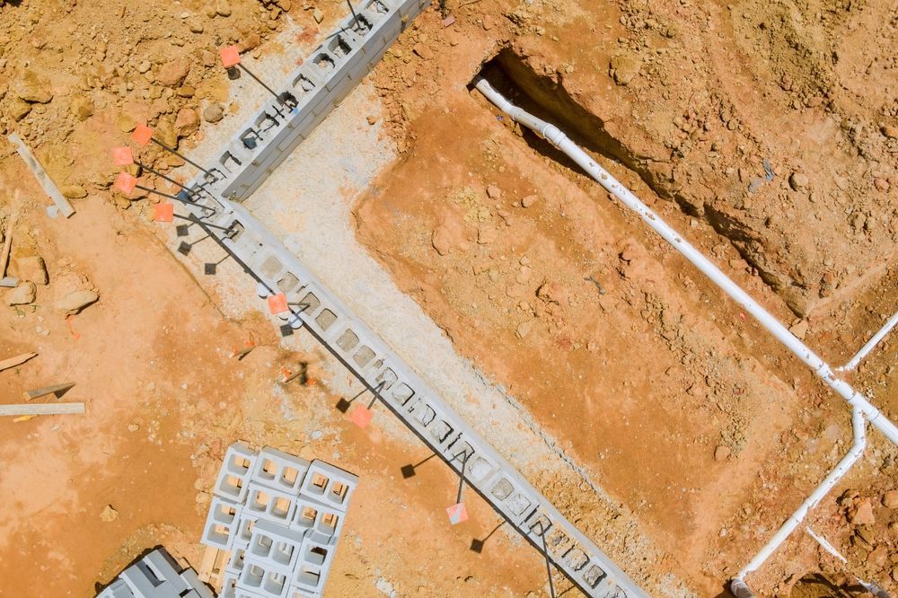 Aerial view of a residential foundation under construction, featuring concrete blocks and white pipes in orange soil.
