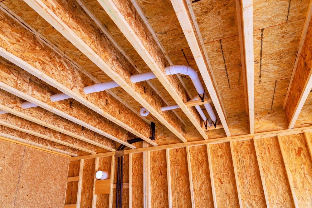 Unfinished wood-frame ceiling with exposed joists, sheathing, and white PVC plumbing pipes running between the beams.