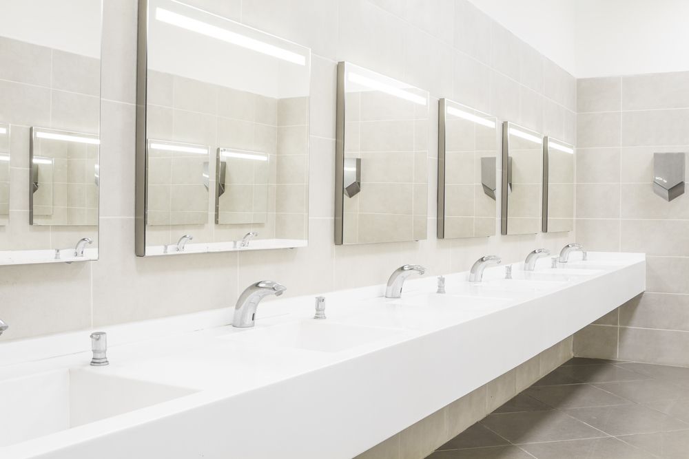 Modern public restroom with a long white communal sink, multiple chrome faucets, and square mirrors on a tiled wall.