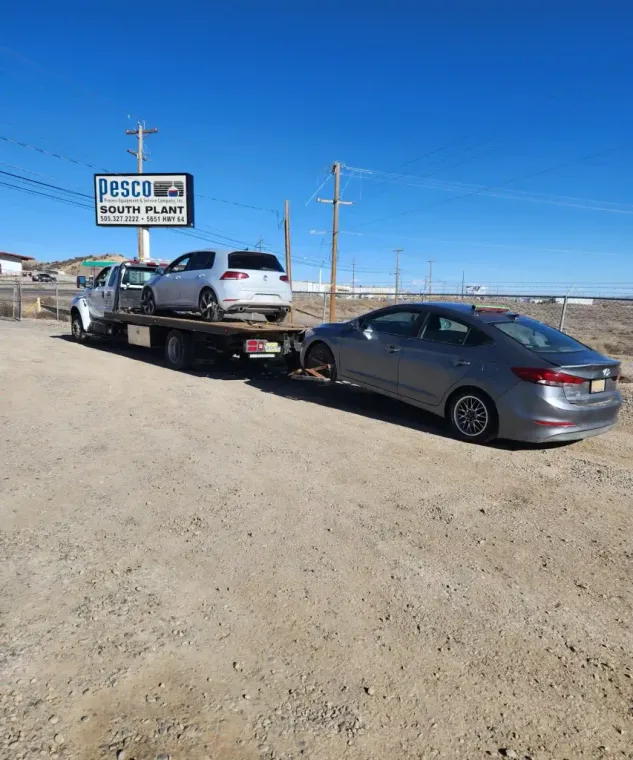 Tow truck carrying a white SUV, with a gray sedan hitched behind, parked on a dirt lot under a blue sky