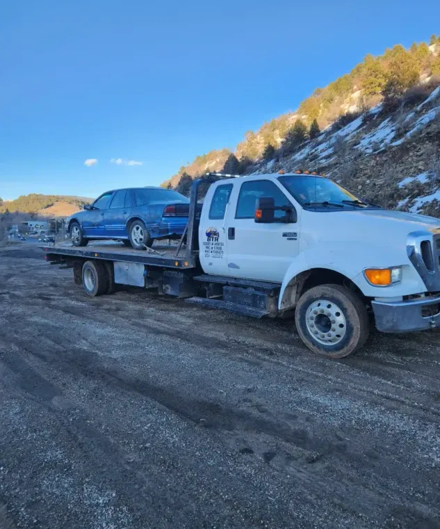 White flatbed tow truck carrying a blue car on a snowy roadside under a clear sky