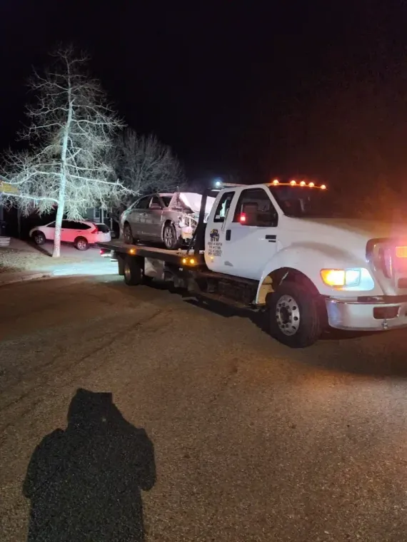 Tow truck with a damaged car loaded on the flatbed at night on a residential street.