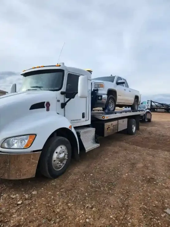 White tow truck carrying a silver pickup on a flatbed in a muddy lot