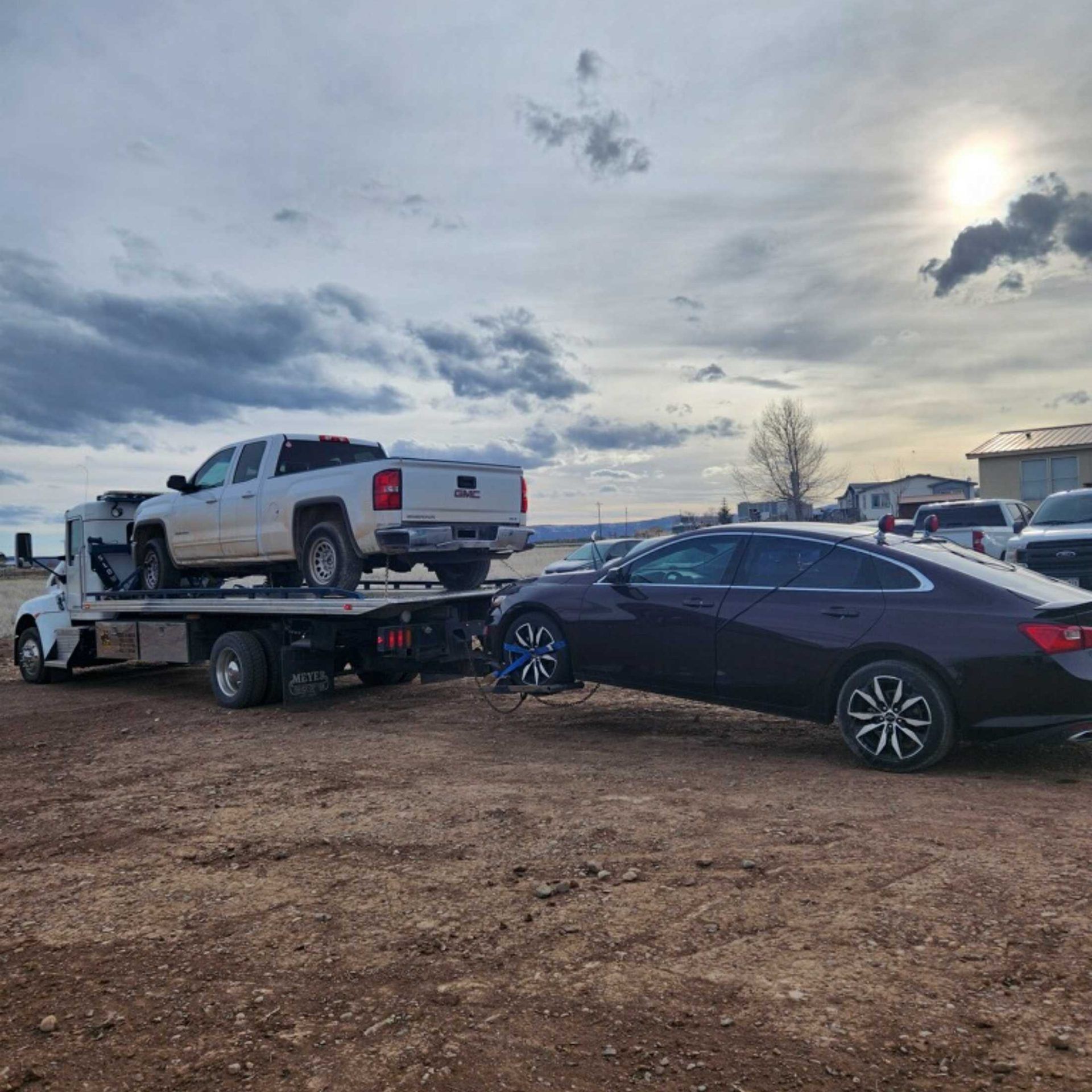 Tow truck hauling a white pickup beside a black sedan in a muddy lot under a cloudy sky