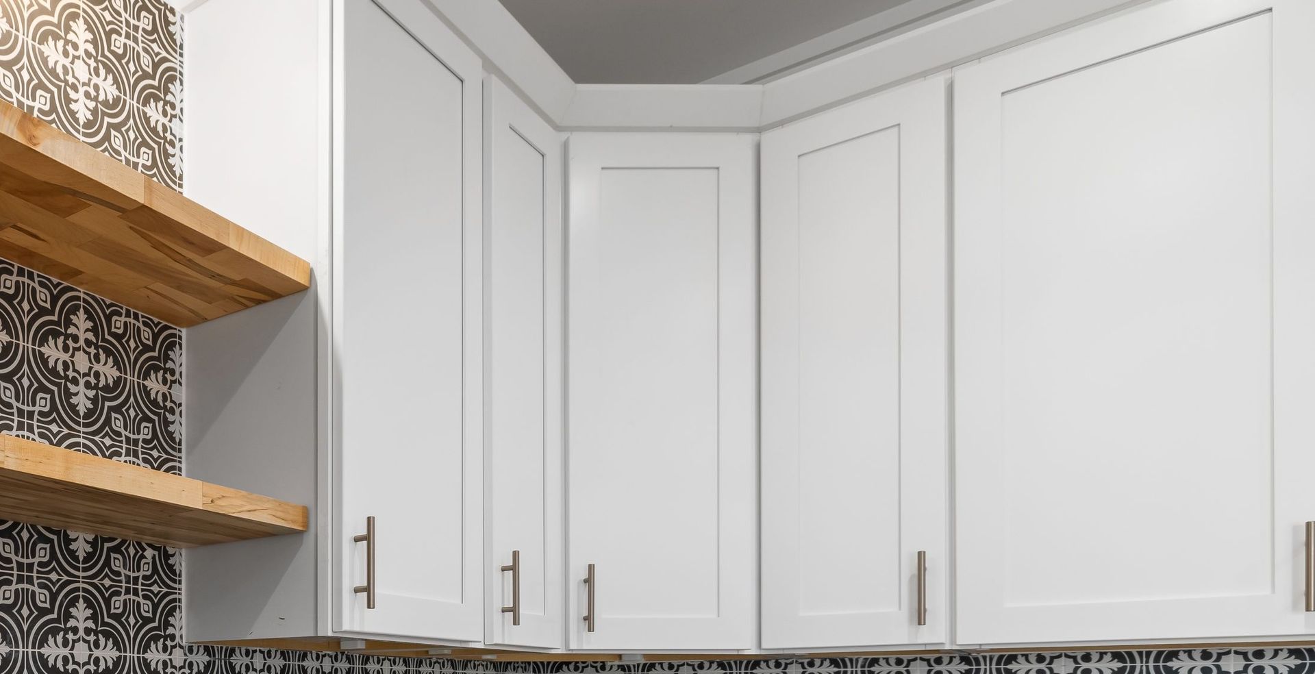 White kitchen cabinets with brass handles beside open wooden shelves and a patterned tile backsplash.