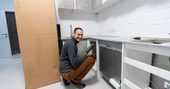 Man kneeling under a kitchen counter, using a drill during cabinet installation.
