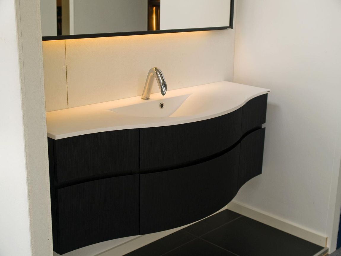 Modern bathroom vanity with white sink, chrome faucet, and black curved cabinets beside a mirror.
