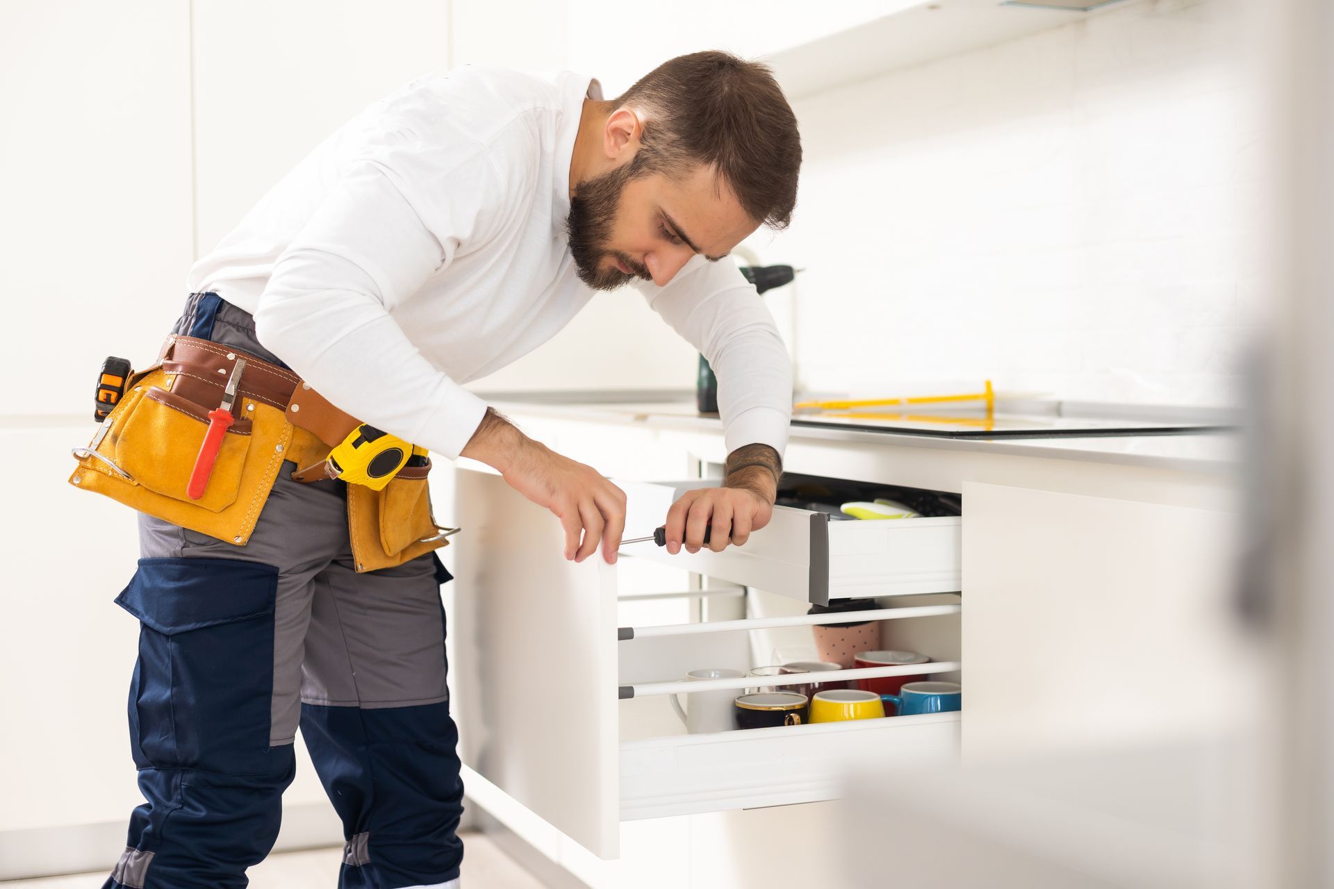 Technician in white shirt and tool belt leaning over an open drawer in a bright kitchen renovation.