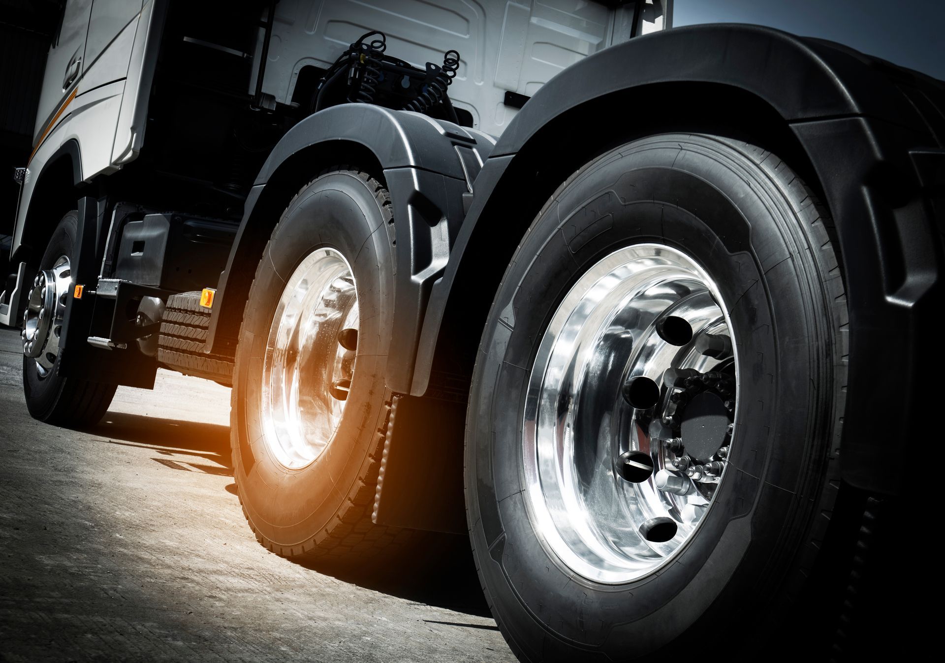 Close-up of a black semi-truck’s chrome wheels on a road, with sunlight reflecting off the tires.