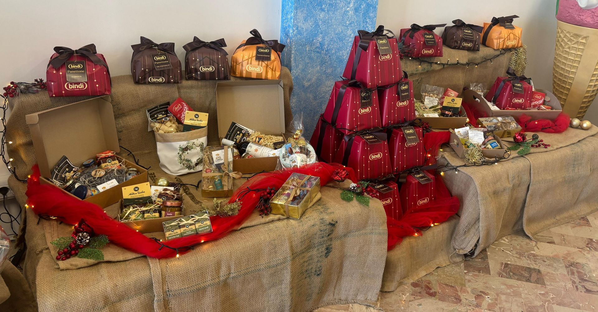 Una composizione di scatole regalo natalizie e sacchetti di cioccolatini disposti su tavoli a più livelli ricoperti da una tela di iuta color beige.
