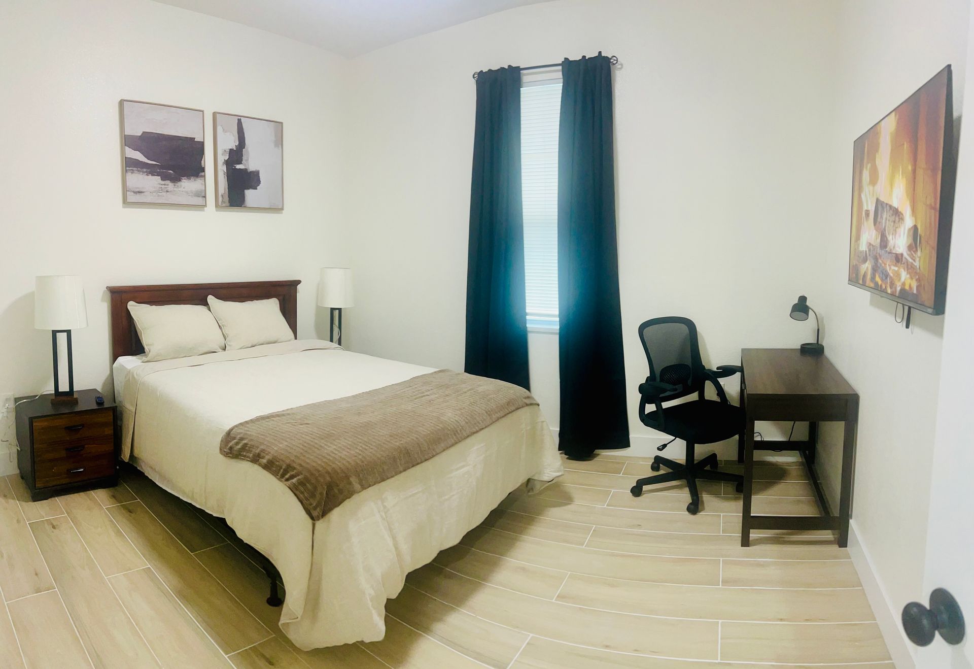 Bedroom with bed, desk, artwork, and window with dark curtains. Light wood floors and white walls.