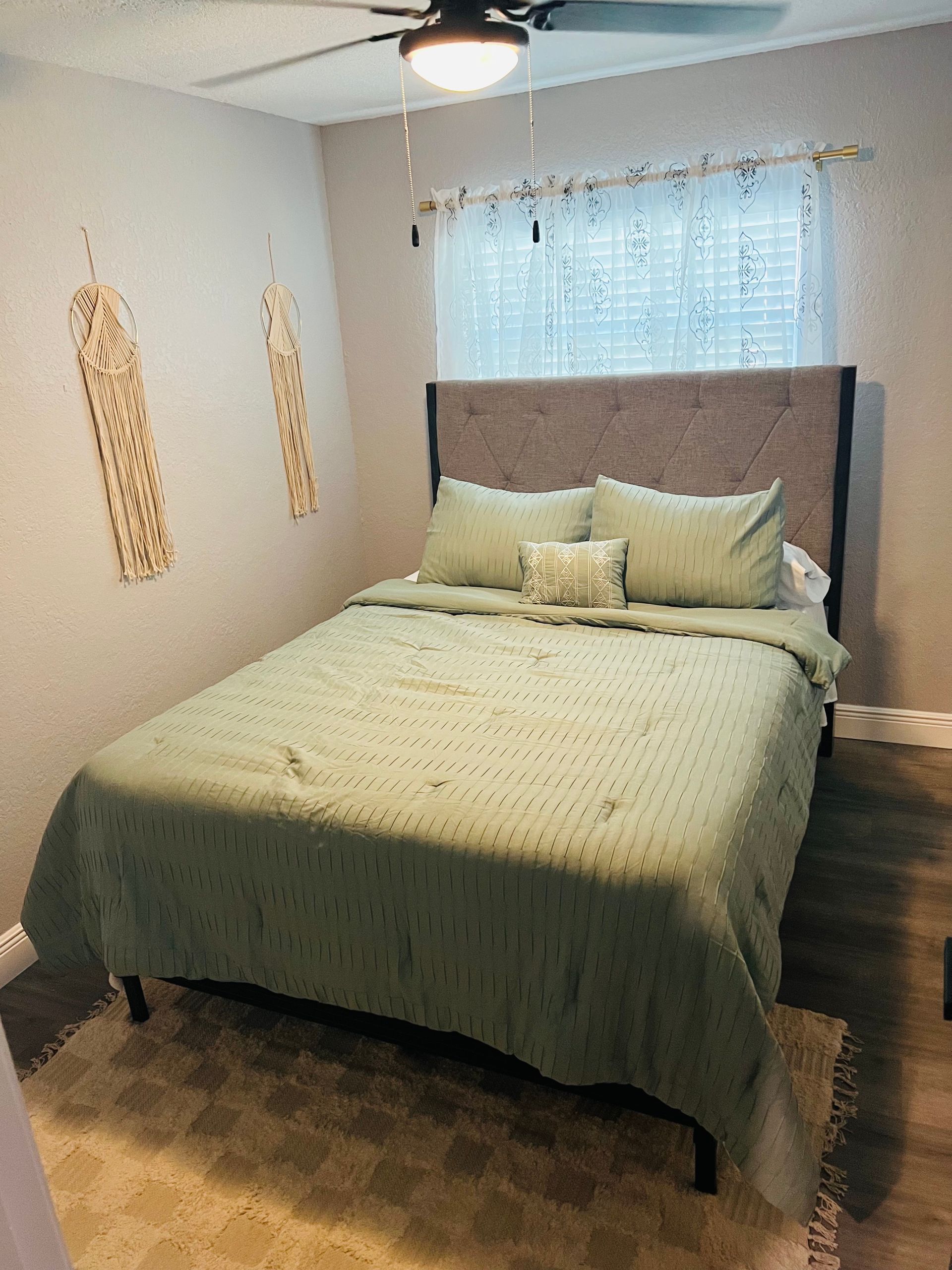 Bedroom with a bed, green bedding, and a window with sheer curtains. Wooden floors and decorative wall art.